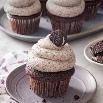 Pinterest graphic of a cupcake topped with Oreo frosting and a mini oreo on a small plate. More Oreo frosted cupcakes in the background.