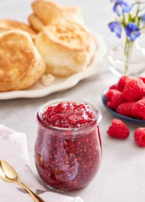A jar of raspberry jam. A platter of biscuits in the back along with a plate of raspberries and some flowers.