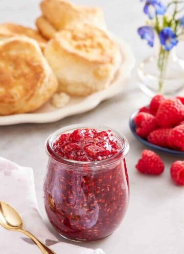 A jar of raspberry jam. A platter of biscuits in the back along with a plate of raspberries and some flowers.