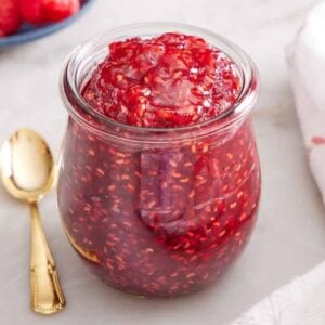 A jar of raspberry jam with a spoon on the side. Raspberries in the background.