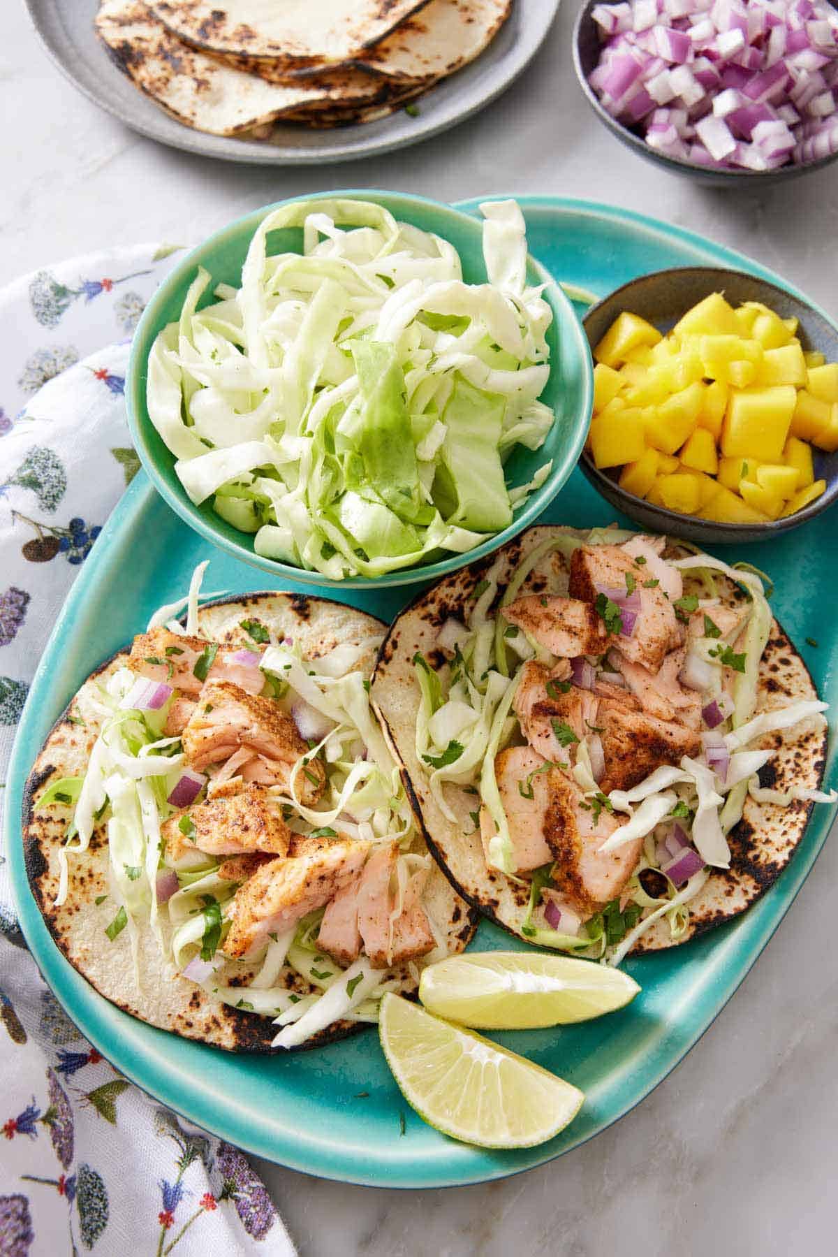 A platter with two salmon tacos with a bowl of cabbage, mango, and cut limes.