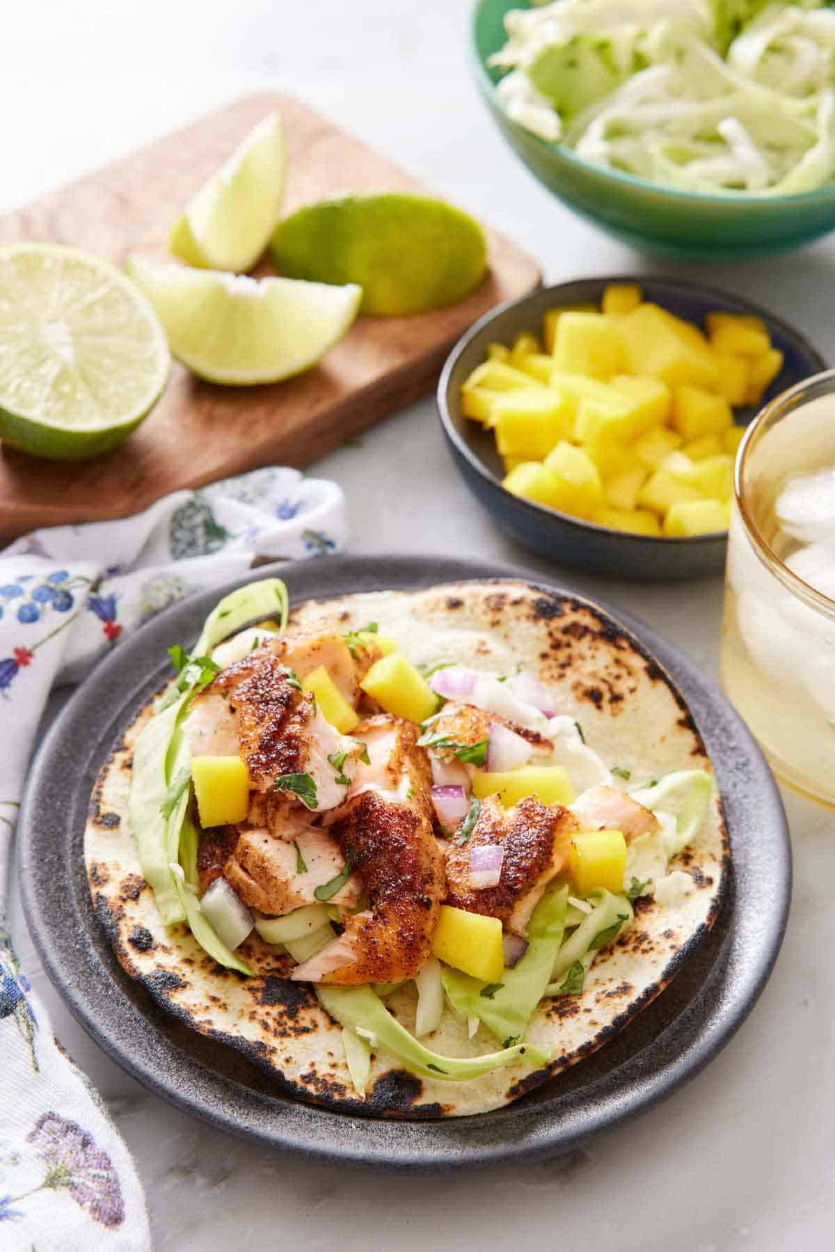 A salmon taco on a plate. A bowl of mangos, cabbage, a drink, and some cut limes in the background.