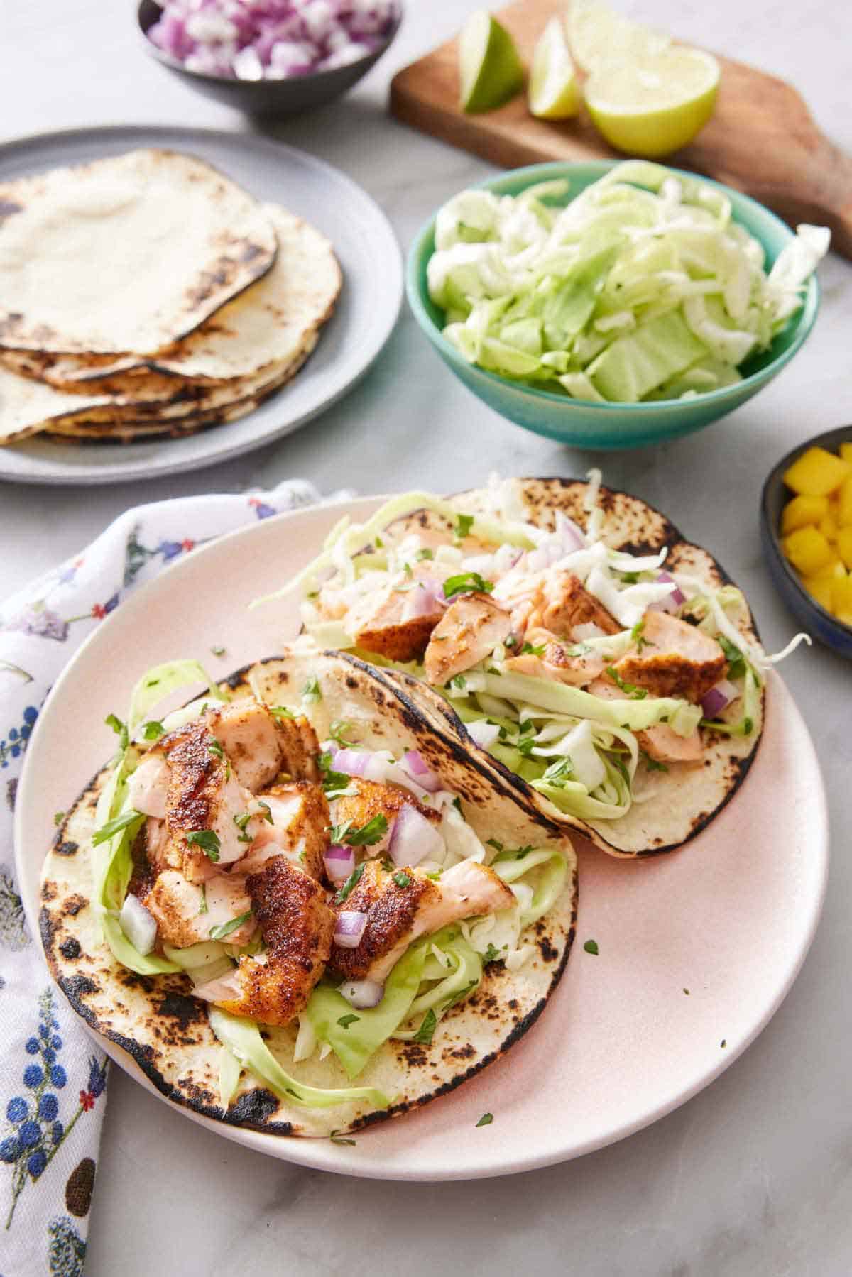 A plate with two salmon tacos. A plate with tortillas, a bowl of cabbage, cut limes, and red onions in the background.