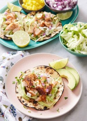 A plate with an open faced salmon taco with a lime wedge and avocado on the side. A platter with more salmon tacos in the background with bowls of cabbage, red onions, and mango.