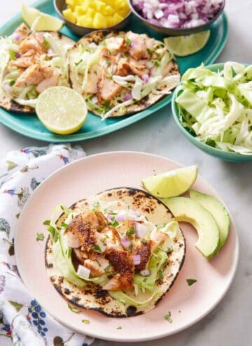 A plate with an open faced salmon taco with a lime wedge and avocado on the side. A platter with more salmon tacos in the background with bowls of cabbage, red onions, and mango.