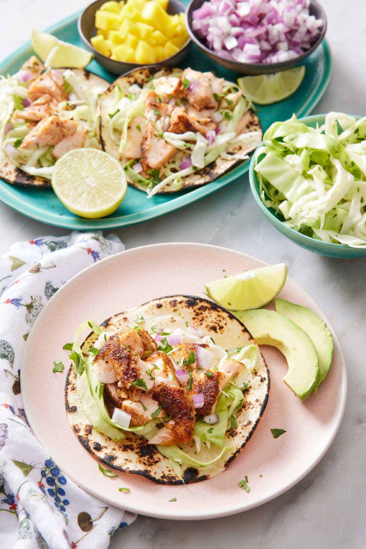 A plate with an open faced salmon taco with a lime wedge and avocado on the side. A platter with more salmon tacos in the background with bowls of cabbage, red onions, and mango.