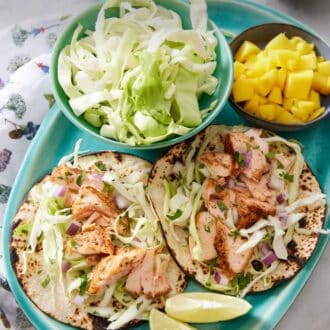 Pinterest graphic of a platter with two salmon tacos with a bowl of cabbage, mango, and cut limes.