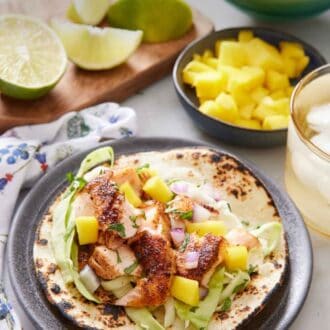 Pinterest graphic of a salmon taco on a plate. A bowl of mangos, a drink, cabbage, and some cut limes in the background.