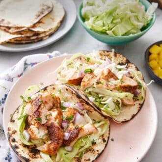 Pinterest graphic of a plate with two salmon tacos. A plate with tortillas, a bowl of cabbage, cut limes, and red onions in the back.