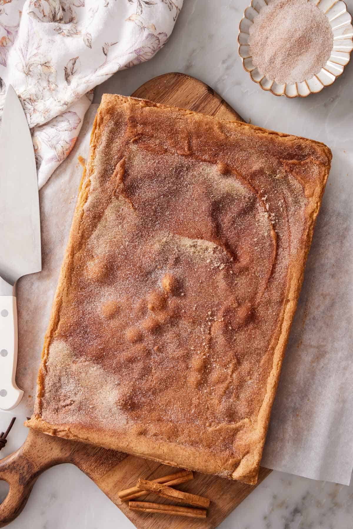 Overhead view of a slab of snickerdoodle bars before slicing on a wooden serving board. Cinnamon sugar off to the side.