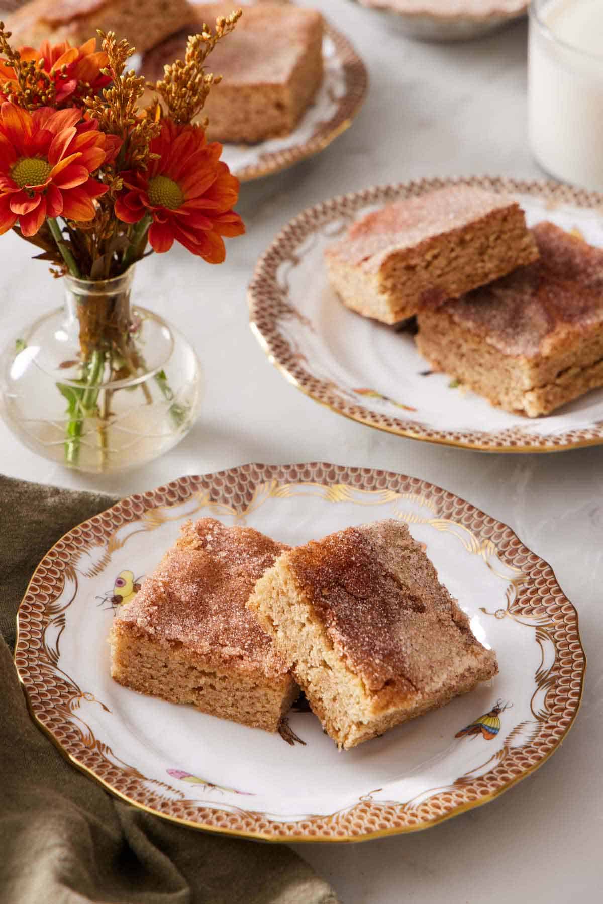 A plate with two pieces of snickerdoodle bars. Another plate with two more bars in the background with a small vase of flowers.