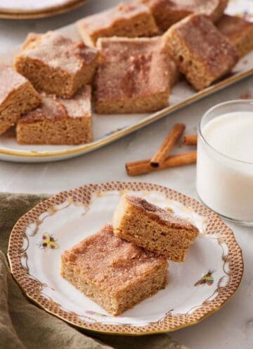 A plate with two pieces of snickerdoodle bars. A glass of milk and platter of more snickerdoodle bars in the background.