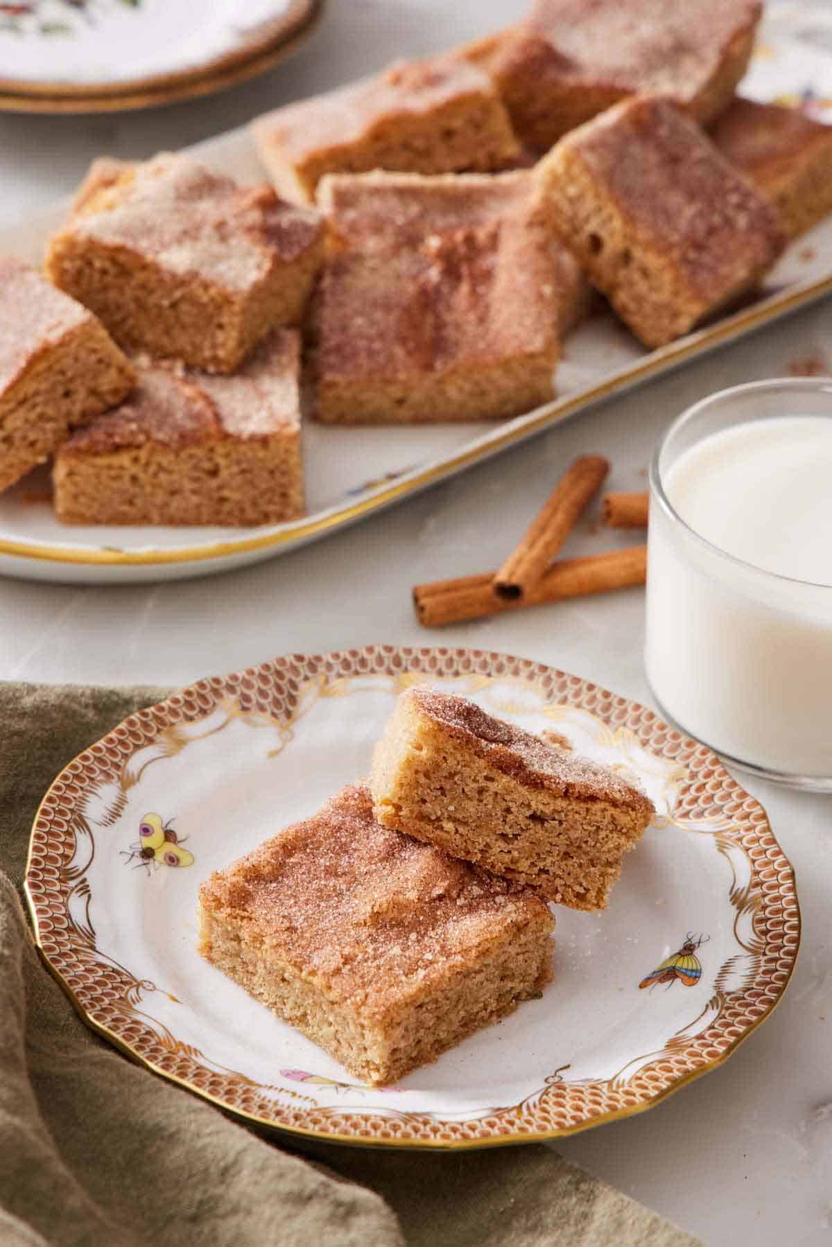 A plate with two pieces of snickerdoodle bars. A glass of milk and platter of more snickerdoodle bars in the background.