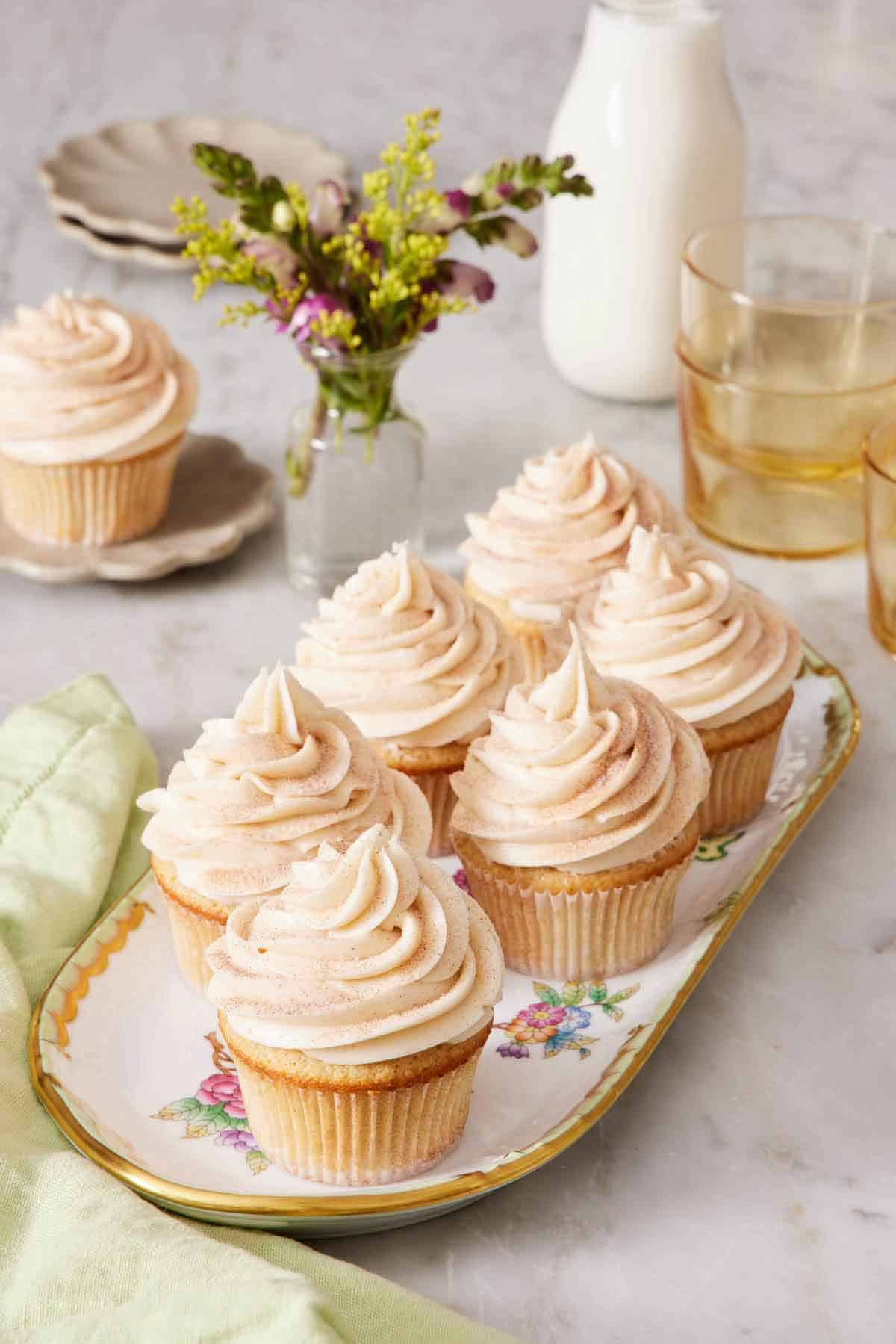 A platter of six snickerdoodle cupcakes. Flowers, drink, milk, and a single snickerdoodle cupcake in the background.