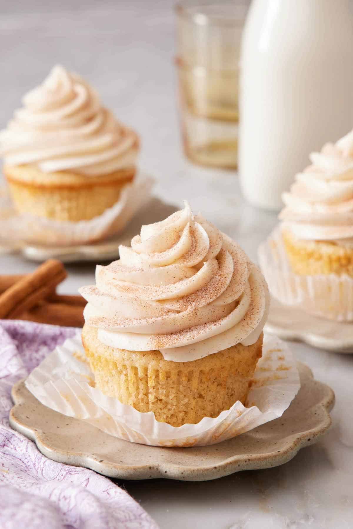 A snickerdoodle cupcake with its liner peeled down on a small plate with two more cupcakes in the background.