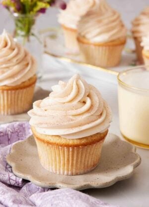 A scalloped plate with a snickerdoodle cupcake on top. A glass of milk in the background along with more cupcakes.