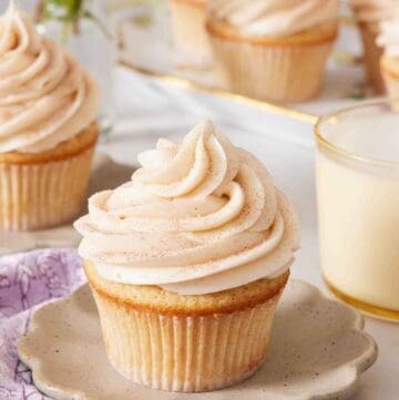 A scalloped plate with a snickerdoodle cupcake on top. A glass of milk in the background along with more cupcakes.