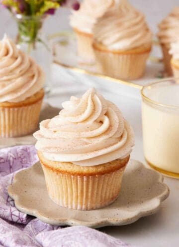 A scalloped plate with a snickerdoodle cupcake on top. A glass of milk in the background along with more cupcakes.