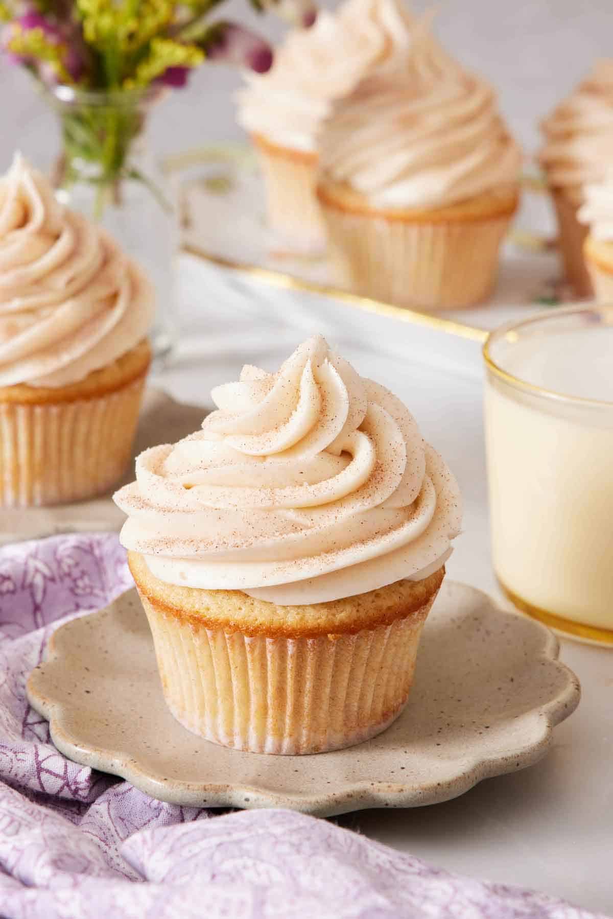 A scalloped plate with a snickerdoodle cupcake on top. A glass of milk in the background along with more cupcakes.