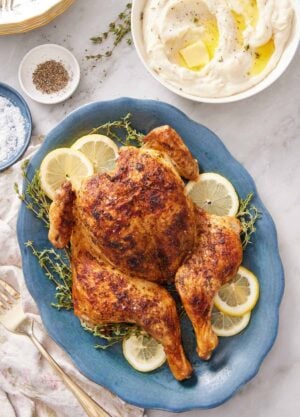 An overhead view of a blue platter of spatchcock chicken with lemon slices and fresh thyme. A bowl of mashed potatoes on the side with salt and pepper.