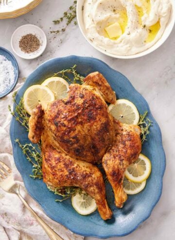 An overhead view of a blue platter of spatchcock chicken with lemon slices and fresh thyme. A bowl of mashed potatoes on the side with salt and pepper.