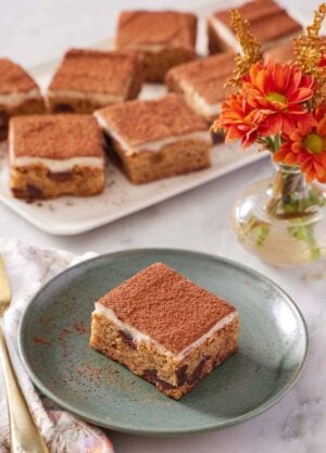 A green plate with a piece of tiramisu blondie. Flowers and a platter of more tiramisu blondies in the background.