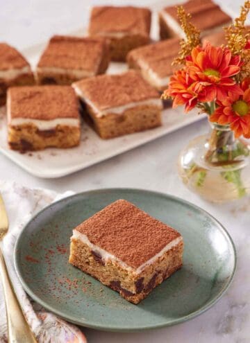 A green plate with a piece of tiramisu blondie. Flowers and a platter of more tiramisu blondies in the background.