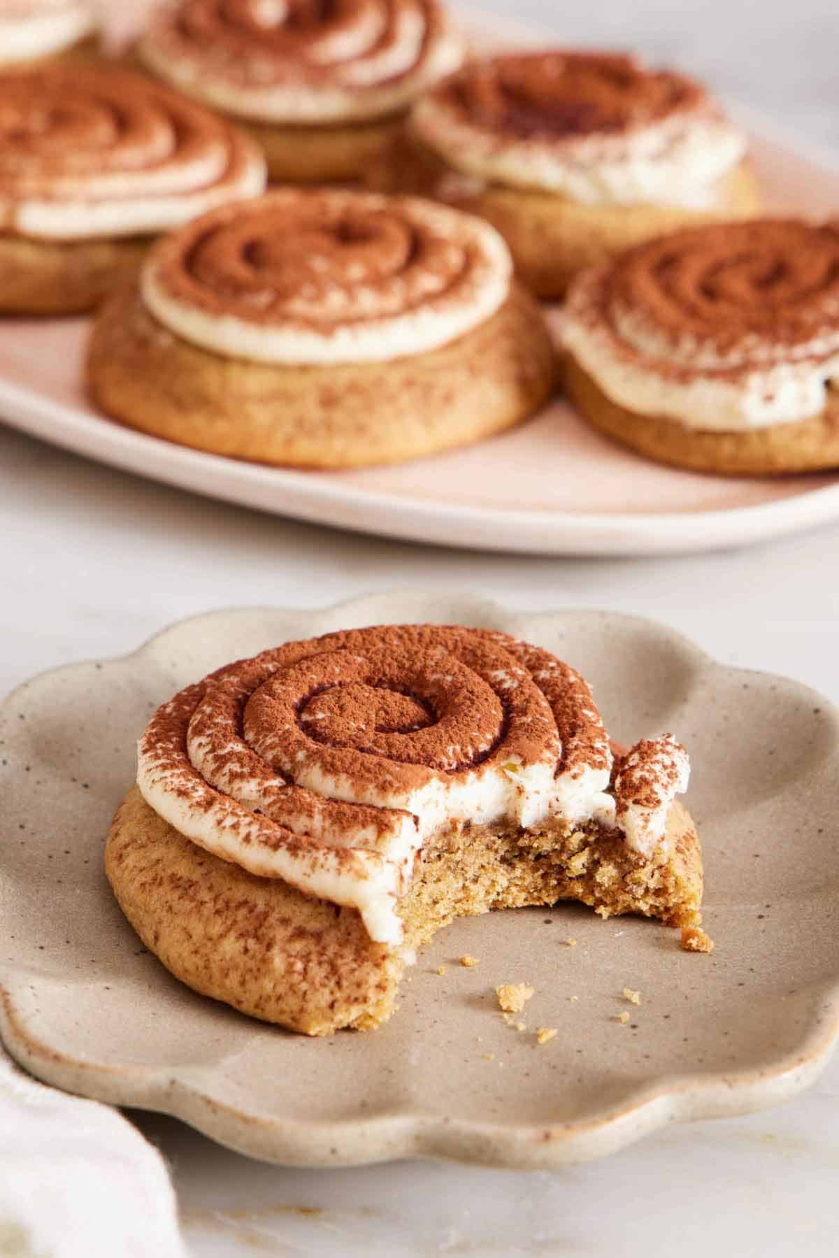 A plate with a tiramisu cookie with a bite taken out. A platter of more cookies in the background.