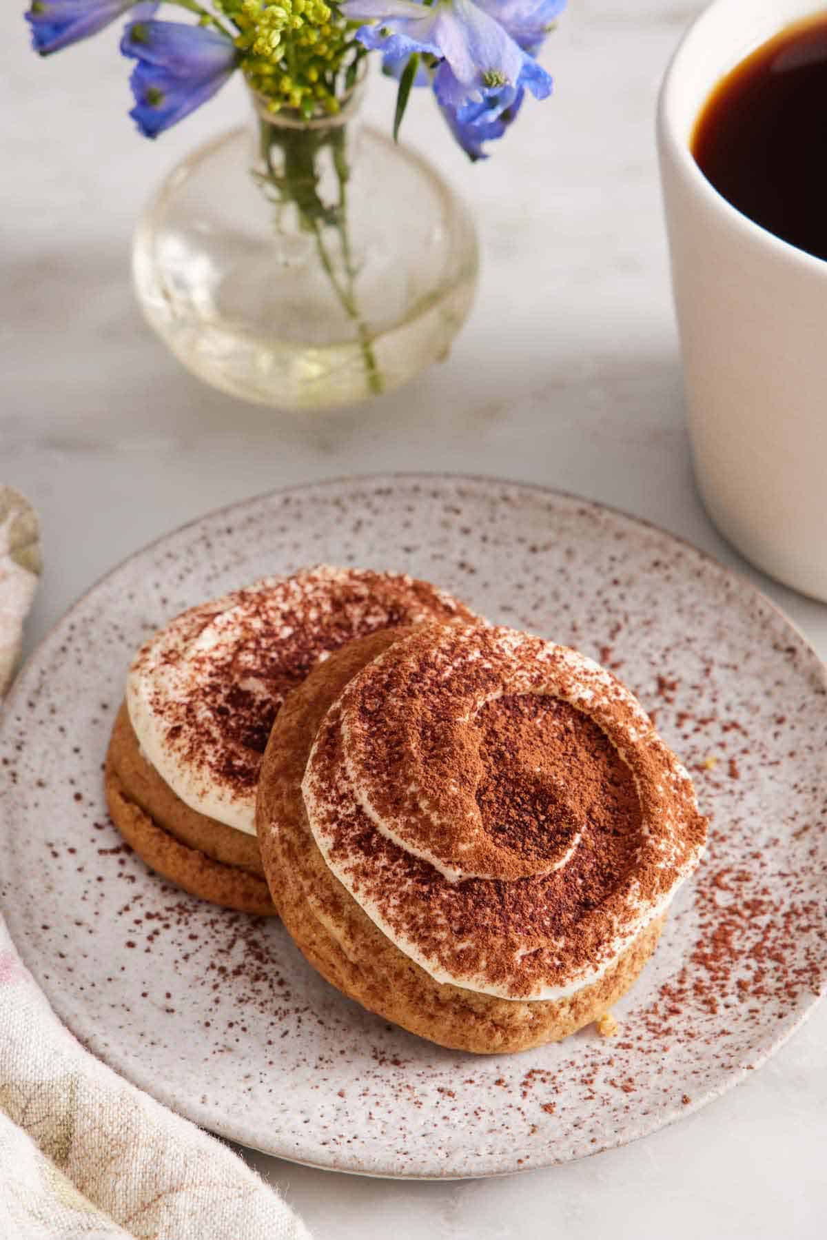 A plate with two tiramisu cookies with a mug of coffee and flowers in the background.