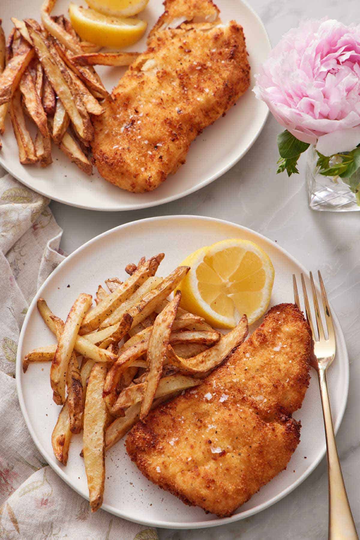 Overhead view of two plated servings of air fryer fish and chips with lemon wedges. a fork on one plate and a flower in a vase on the side.