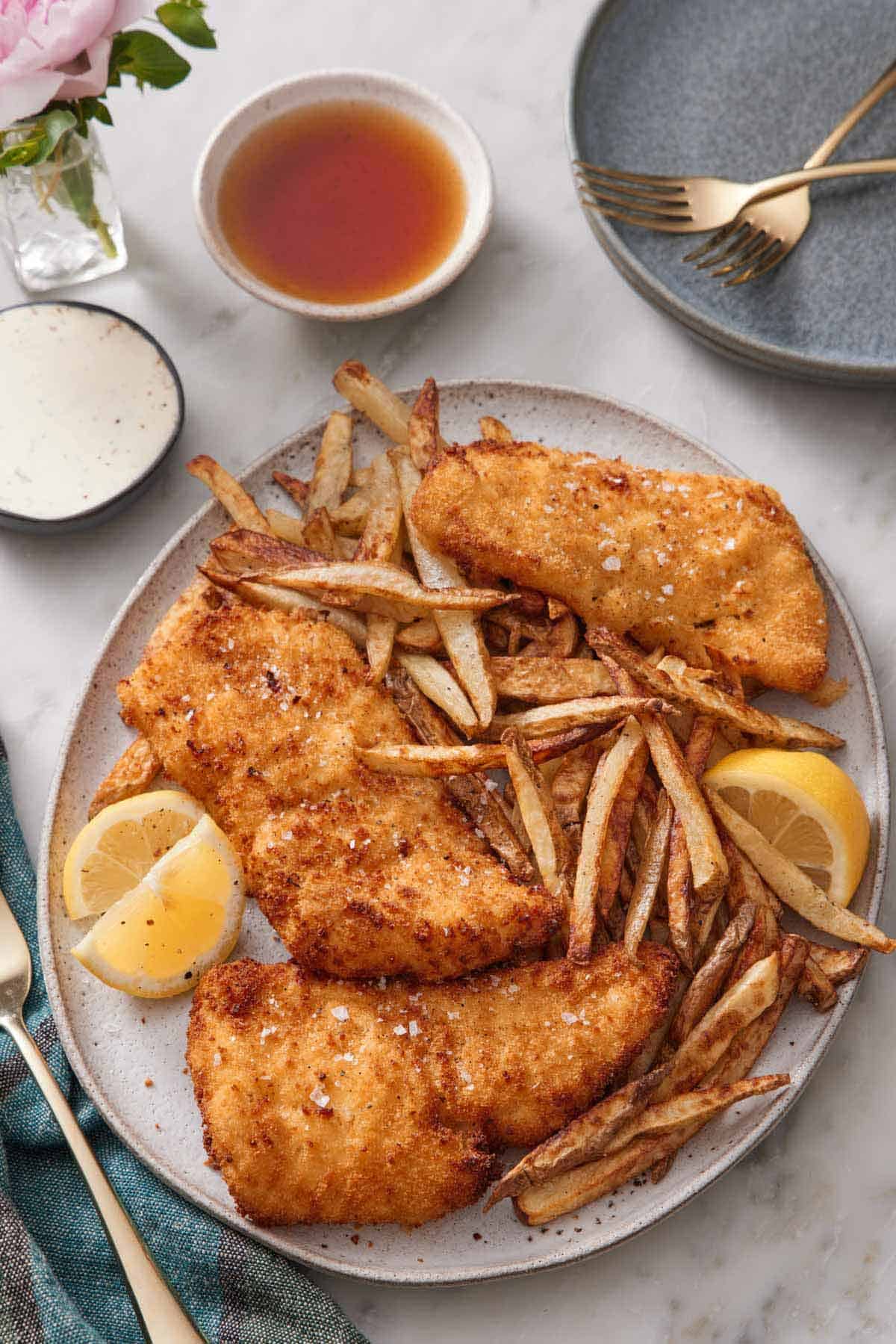 Overhead view of a platter of air fryer fish and chips with three lemon wedges topped with flaky salt. A bowl of tartar sauce, malt vinegar, and stack of plates with forks on the side.