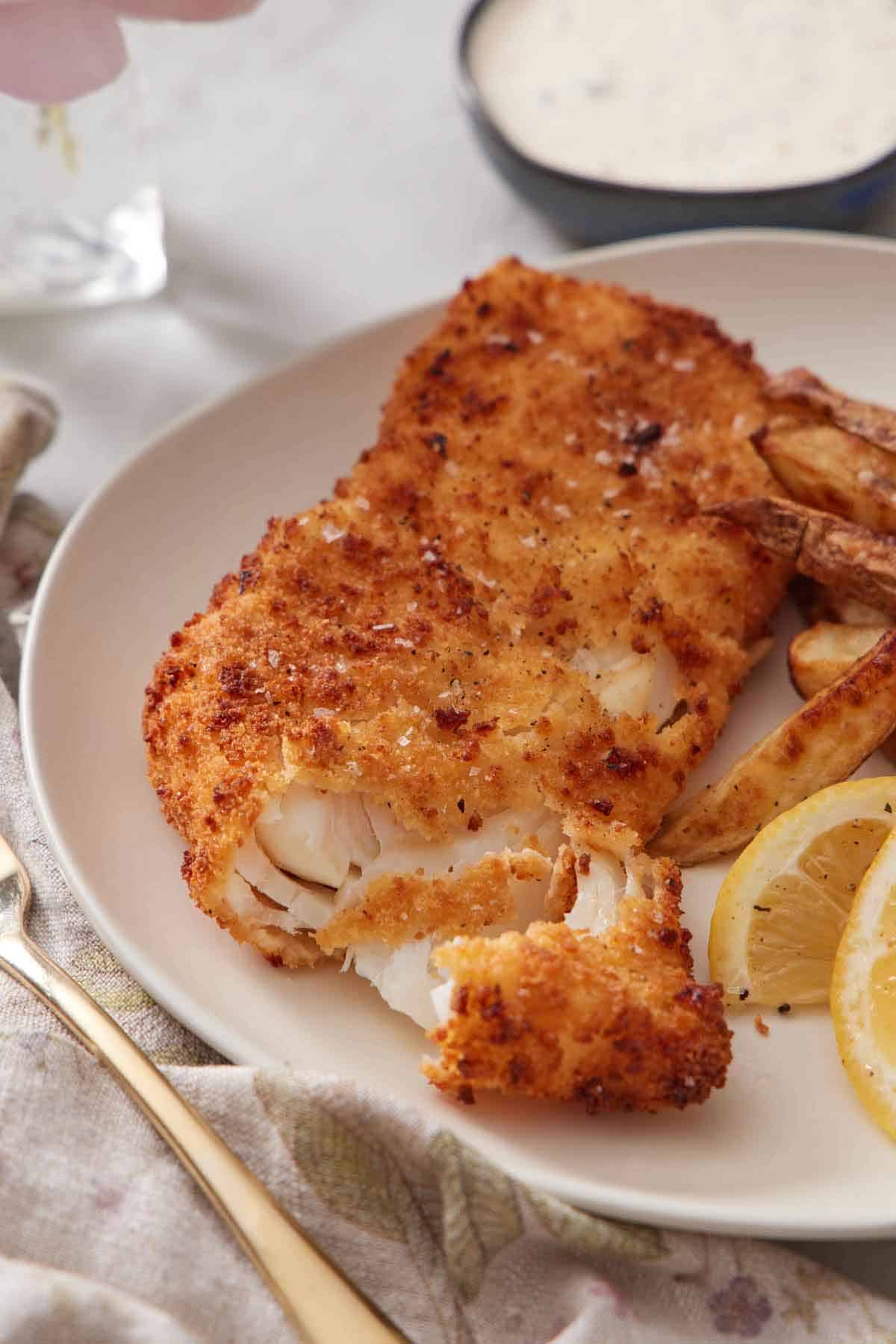 A close up view of a piece of air fryer fish flaked opened with some fries and lemon wedges on the side.