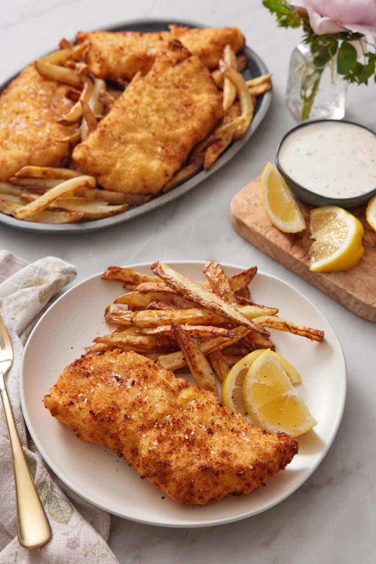 A plate of air fryer fish and chips with lemon wedges. A platter with more in the background also with some lemon wedges and tartar sauce on the side.