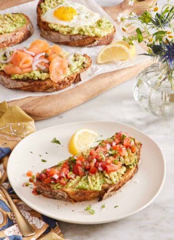 A plate with avocado toast topped with pico de gallo and a lemon wedge on the side. A wooden serving board in the background with three pieces of avocado toast with various toppings and some flowers on the side.