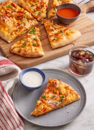 A plate with a slice of buffalo chicken pizza with a bowl of dip. The rest of the pizza in the background with more buffalo sauce on the side and a glass of soda.