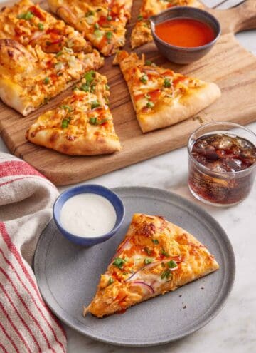 A plate with a slice of buffalo chicken pizza with a bowl of dip. The rest of the pizza in the background with more buffalo sauce on the side and a glass of soda.