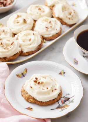 A plate with a carrot cake cookie with a platter in the background with more cookies along with a cup of coffee.