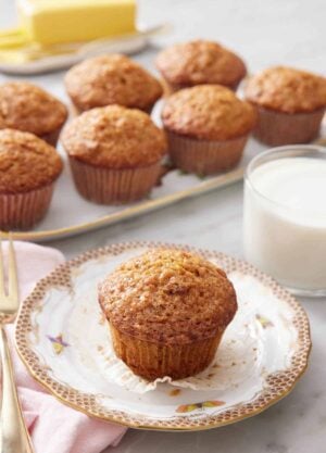 A plate with a carrot muffin with the liner pulled down. A glass of milk in the background with a platter of more muffins.