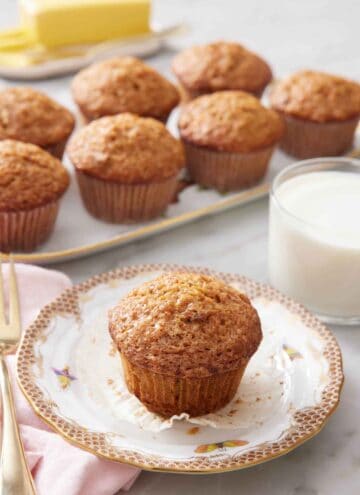 A plate with a carrot muffin with the liner pulled down. A glass of milk in the background with a platter of more muffins.