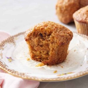A plate with a carrot muffin with a bite taken out. More muffins in the background.