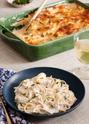 A bowl of chicken tetrazzini with the baking dish of more chicken tetrazzini in the background along with a drink.