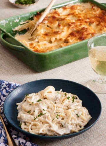 A bowl of chicken tetrazzini with the baking dish of more chicken tetrazzini in the background along with a drink.