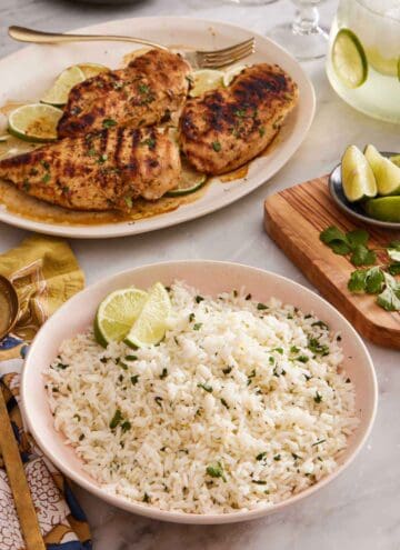 A bowl of cilantro lime rice with grilled chicken in the background. Cut limes and cilantro on the side.