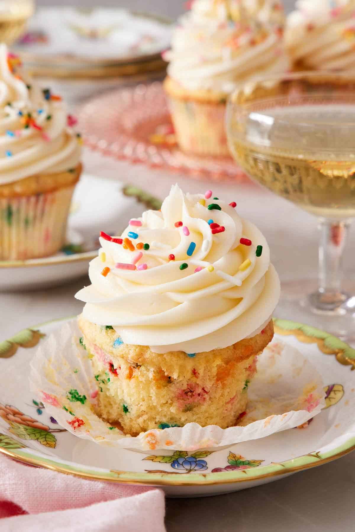 A funfetti cupcake on a plate with the paper liner pulled down. A bubbly drink in the back with more funfetti cupcakes in the background.