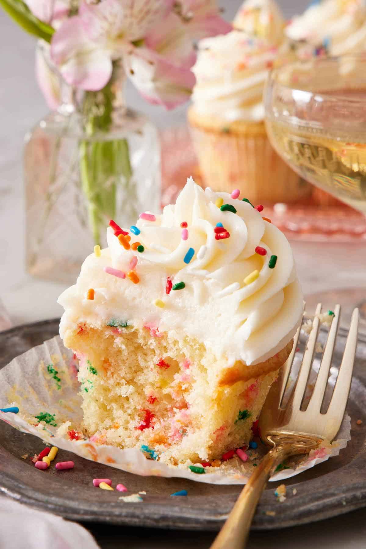 A funfetti cupcake topped with frosting and rainbow sprinkles with a bite taken out and the paper liner pulled down. A fork beside the cupcake. More cupcakes in the background with a vase of flowers.