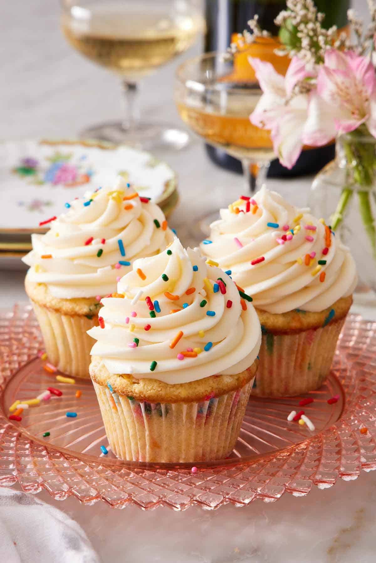 A plate with three funfetti cupcakes with frosting and rainbow sprinkles. Flowers, drinks, and plates in the background.