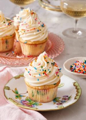 A plate with a funfetti cupcake topped with frosting and rainbow sprinkles. More funfetti cupcakes in the background and a small bowl of sprinkles.