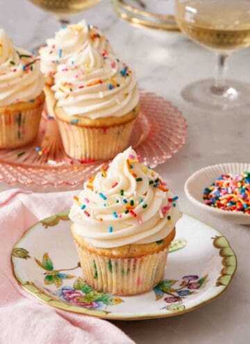 A plate with a funfetti cupcake topped with frosting and rainbow sprinkles. More funfetti cupcakes in the background and a small bowl of sprinkles.