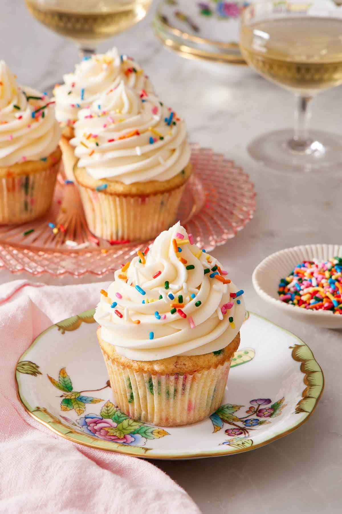 A plate with a funfetti cupcake topped with frosting and rainbow sprinkles. More funfetti cupcakes in the background and a small bowl of sprinkles.