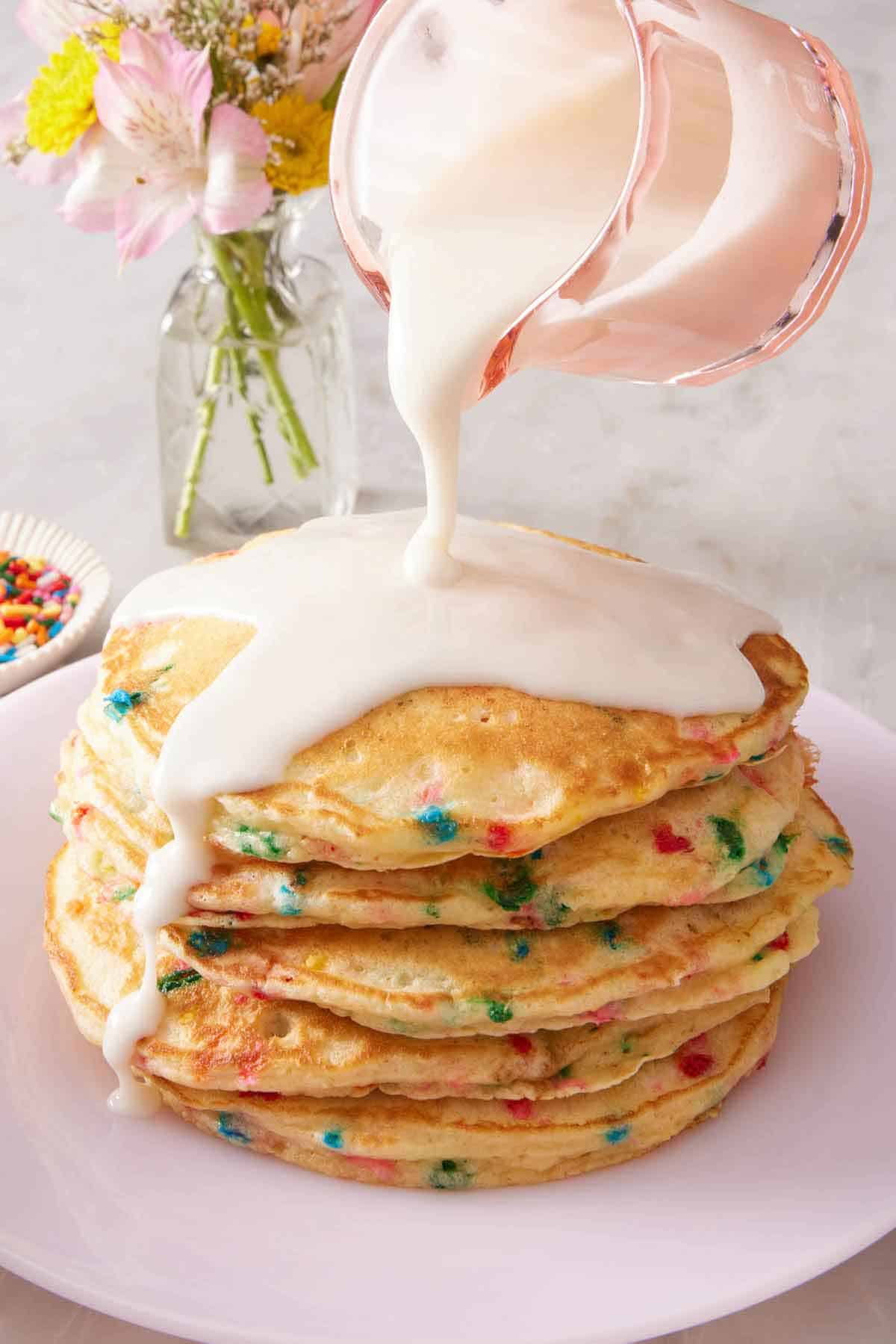 Icing poured over a stack of funfetti pancakes. A small vase of flowers in the background.